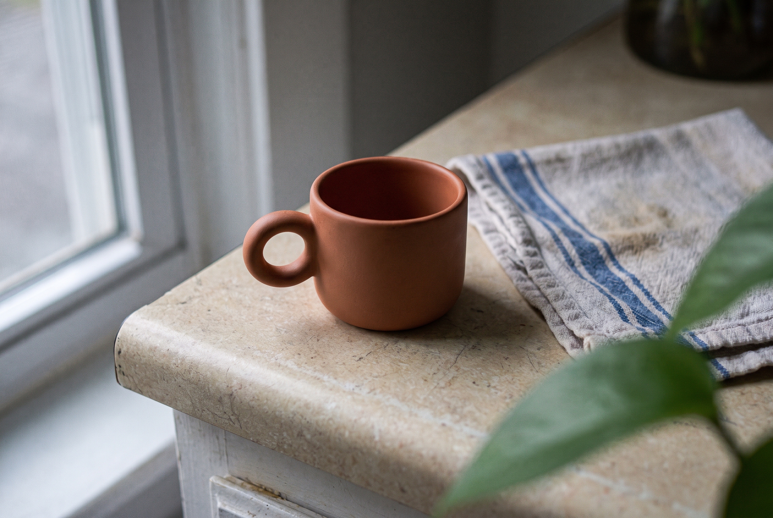 Terracotta ceramic espresso cup on a kitchen counter with a towel and plant leaf in frame